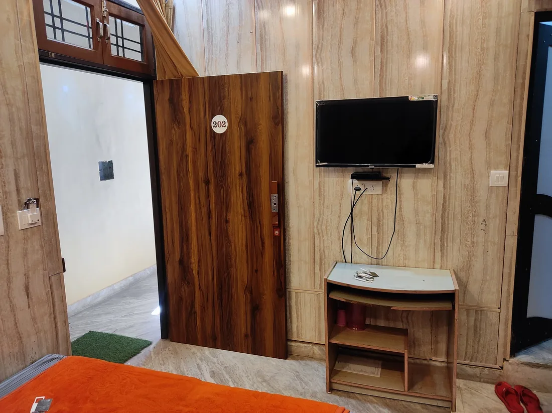 Guest room 202 with wall mounted television, wooden door and orange bedcover at Sri Yoga Ashram