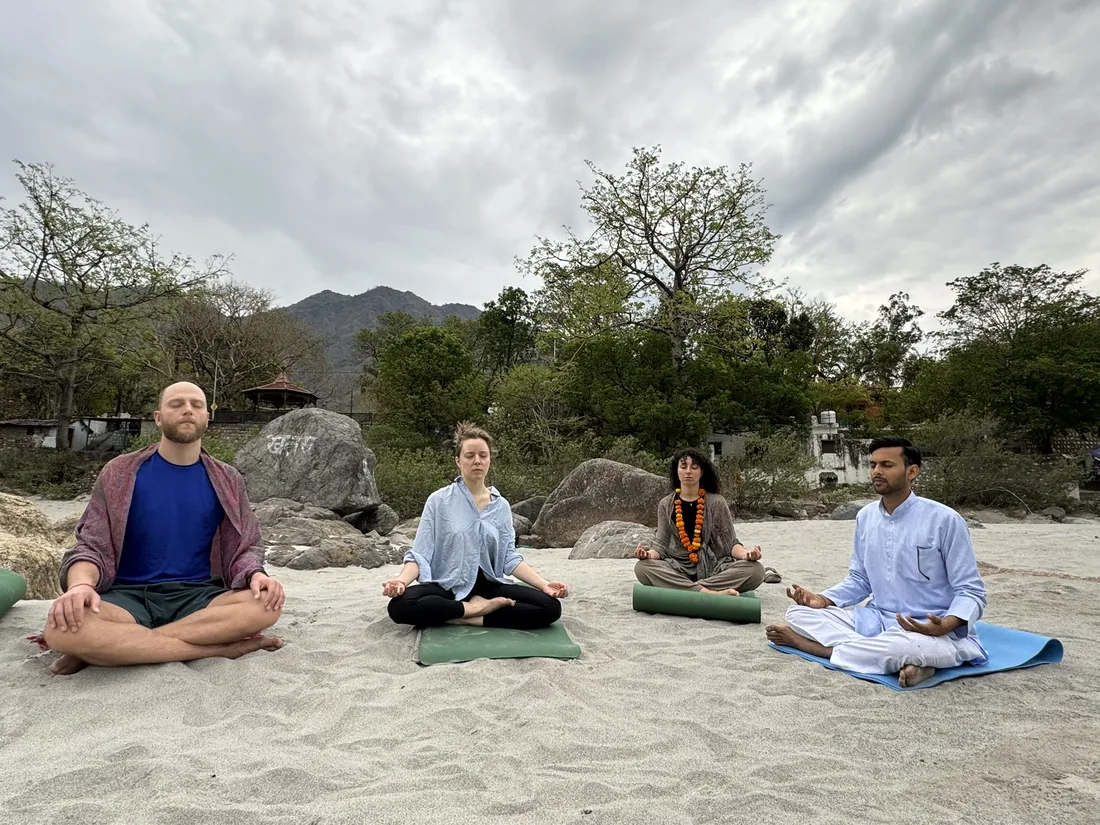 Four students meditating in lotus pose on Ganga sand with Himalayan foothills behind