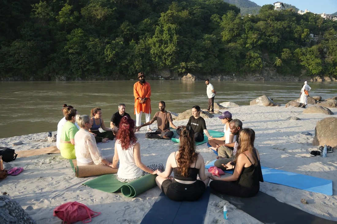 Yoga students meditate in a circle with teacher on Ganges riverbank in Rishikesh