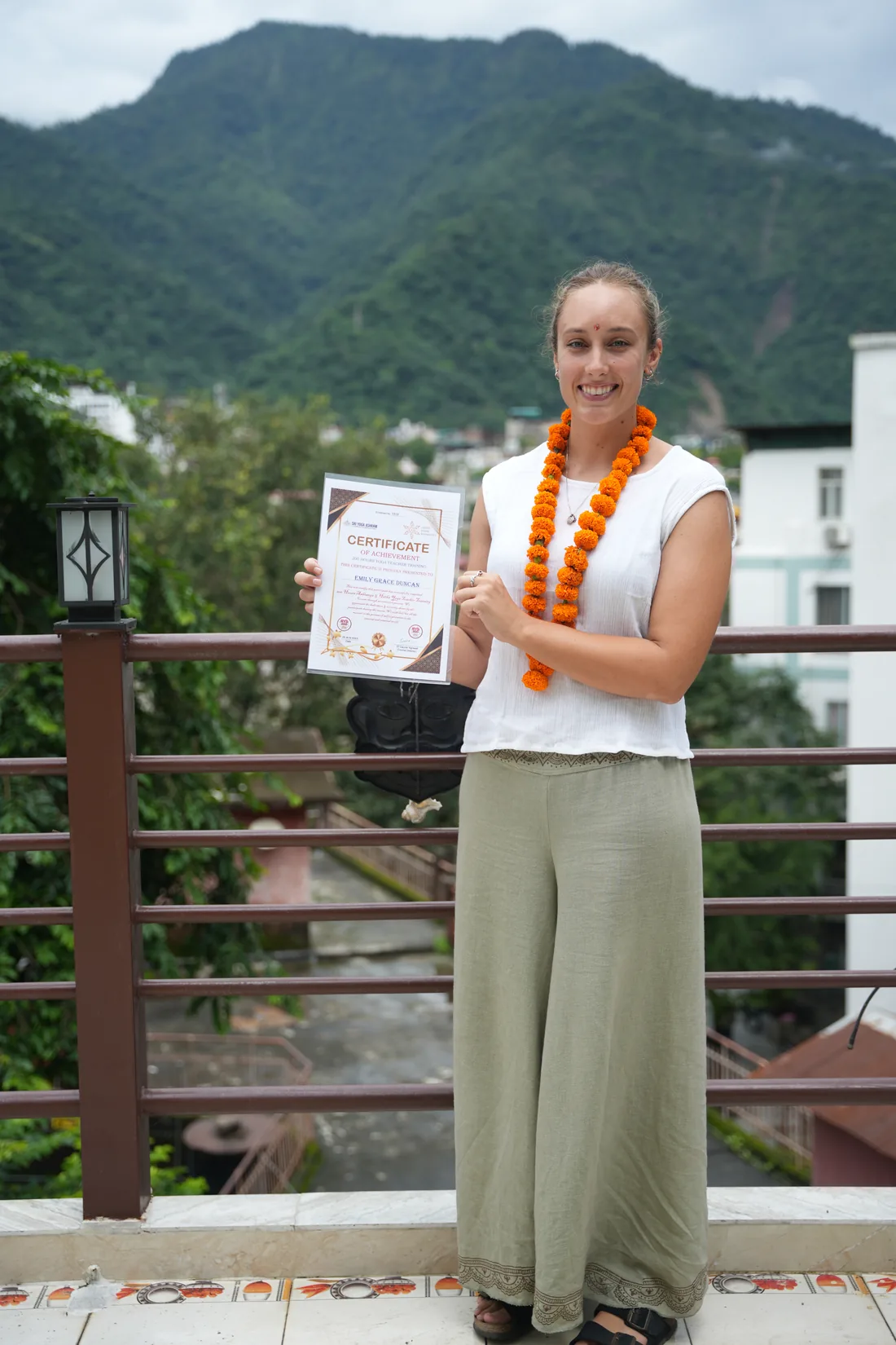 Yoga graduate holding certificate on ashram terrace with green Himalayan foothills behind Rishikesh