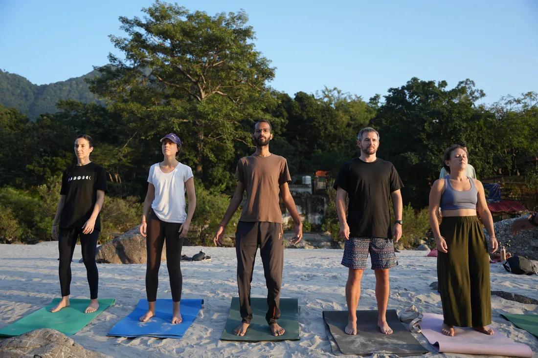 Yoga students standing in Tadasana on the Ganga riverbank at sunrise in Rishikesh
