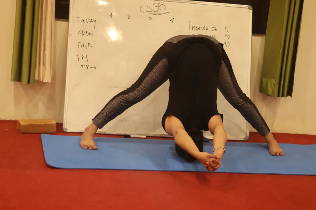 Student in wide legged forward fold beside the class schedule board at Sri Yoga Ashram